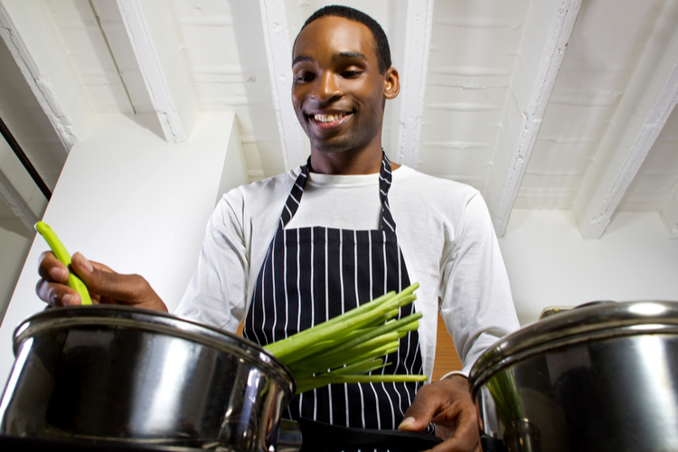 close up of a young black man wearing an apron and cooking at home. he is in a domestic kitchen and preparing a vegetarian meal.