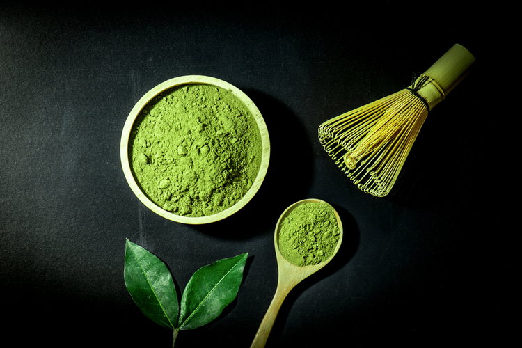 Matcha powder on a spoon and wooden bowl with the matcha leaf and whisk.