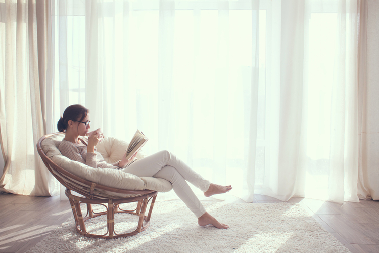 Young woman at home sitting on modern chair in front of window relaxing in her living room reading book and drinking coffee or tea.