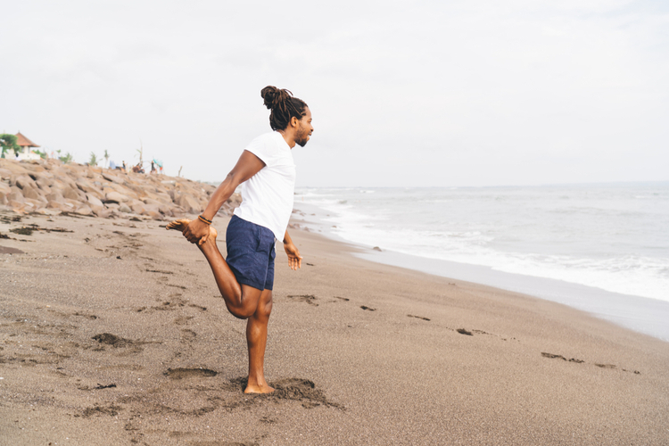 Male in activewear doing stretching exercise for legs while training on beach.