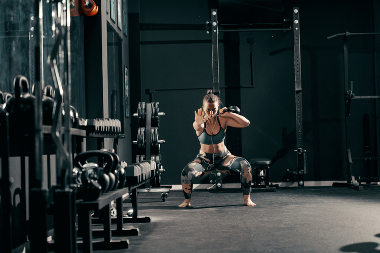Woman with earphones doing workouts with kettlebell in squat position barefoot training.