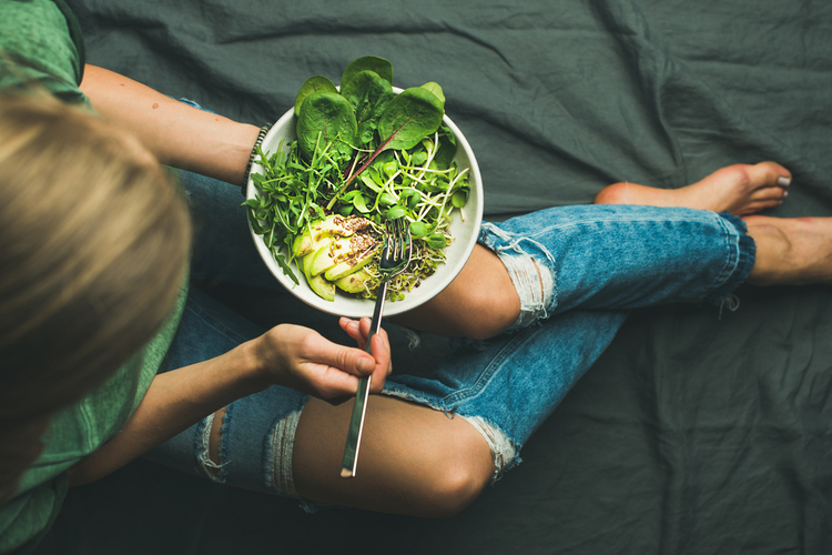 Green vegan breakfast meal in bowl with spinach, arugula, avocado, seeds and sprouts. Girl in jeans holding fork with knees and hands visible, top view. Clean eating, detox, vegetarian food concept
