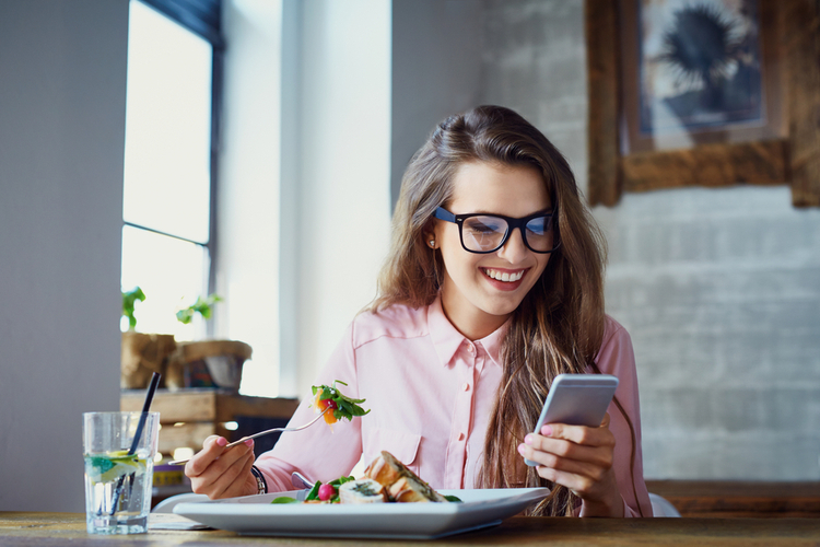 Young woman eating salad at restaurant and texting on smartphone with her diet coach.