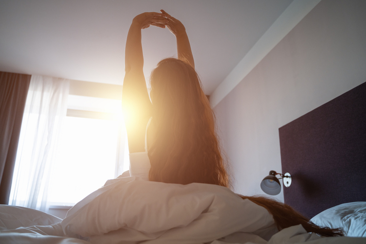 Young woman wakes up, gets up on bed and stretches hands up in morning sunlight.