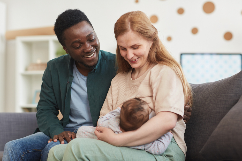 Portrait of happy interracial family sitting on couch at home together with mature mother breastfeeding baby