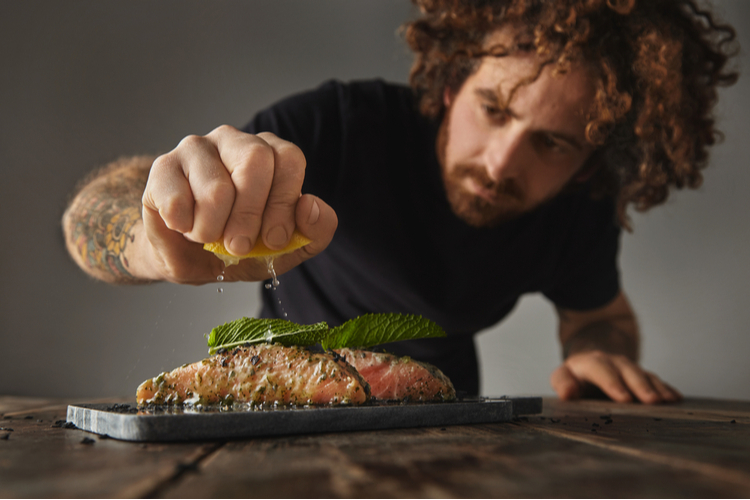 Man cooks healthy meal, squeezes lemon on two raw pieces of salmon decorated with mint leaf.