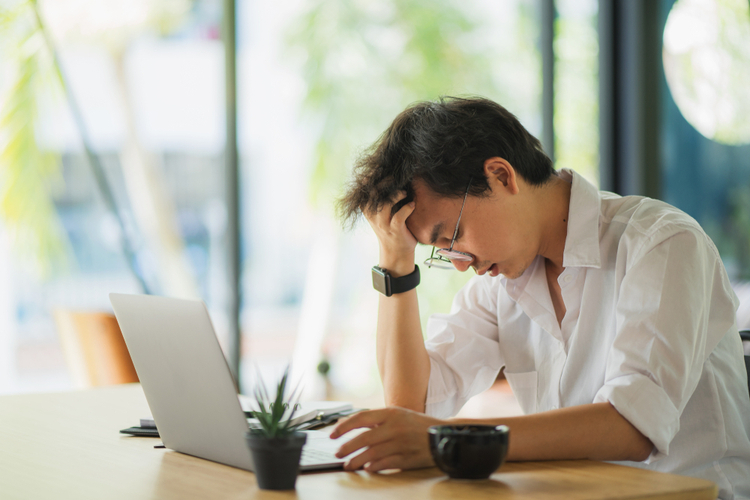 A man in front of his laptop, looking stressed while holding his head. Seems like he's having headache as effect of stress in the body.