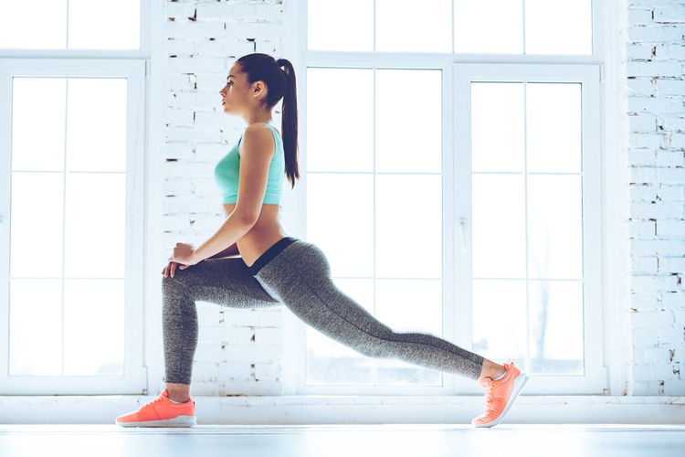 Side view of young beautiful young woman in sportswear doing stretching while standing in front of window at gym.