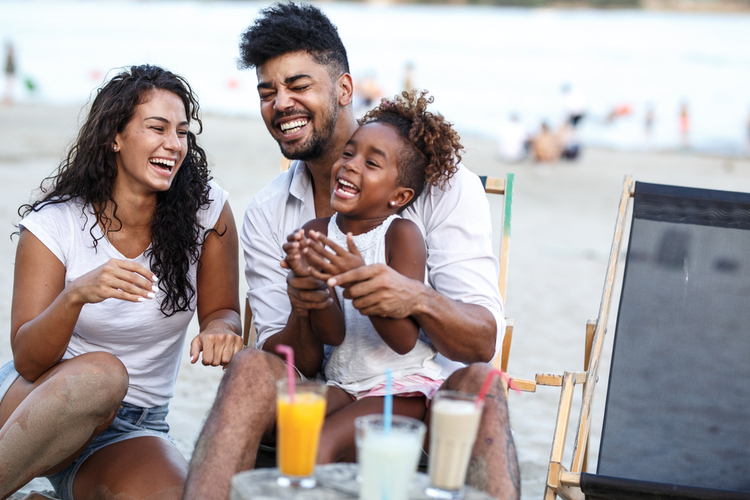 Young mixed race family sitting and relaxing at the beach with summer drinks on beautiful summer day.