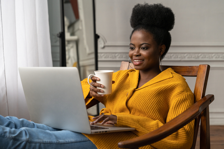 Woman with afro hairstyle wear yellow cardigan having coffee while working on her laptop.