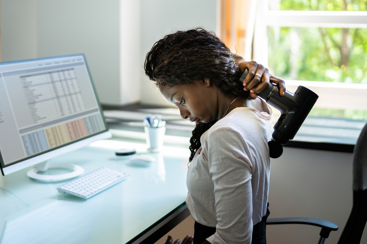 An African American woman using a massage gun for percussive therapy in her office. 