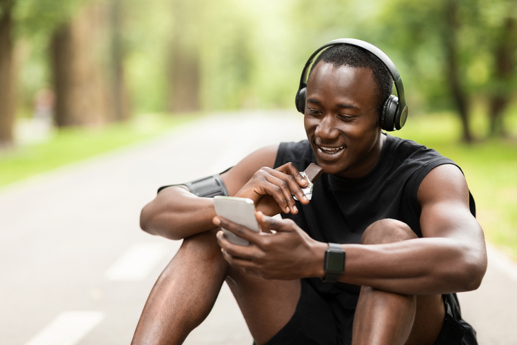 Positive Black Sportsman Eating Protein Bar And Using Phone While Resting, Exercising at Park
