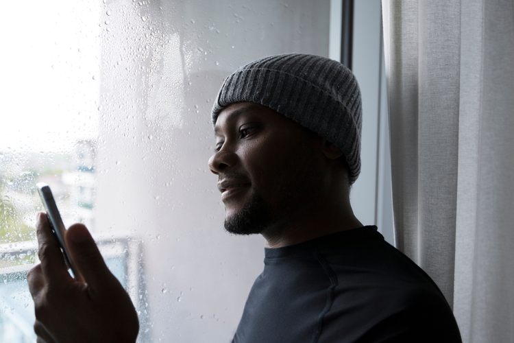 Man smiling and looking at his cellphone while standing by the window on a rainy day.