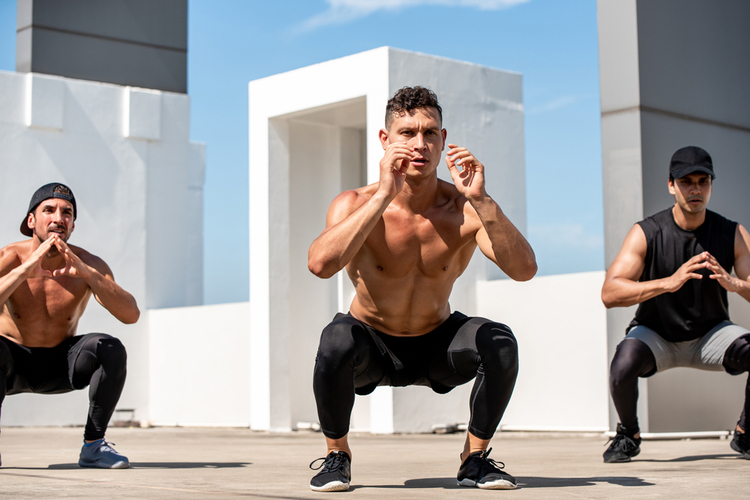 Group of fit sports men doing squat bodyweight workout training outdoors on building rooftop in sunlight 