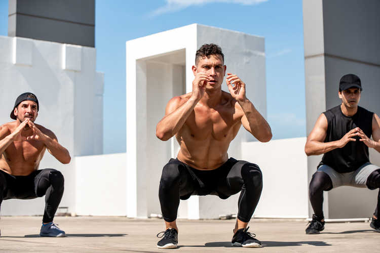 Group of fit sports men doing squat bodyweight workout training outdoors on building rooftop in sunlight