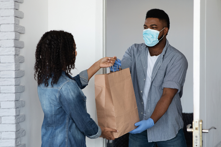 Food delivery during coronavirus. Black courier guy wearing medical mask delivering grocery order to young woman's home