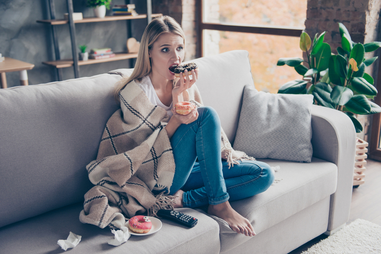Woman sitting on a couch in living room under blanket stress eating donuts.