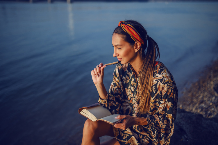 Woman in floral dress and with headband sitting on rock on shore next to river and writing diary.