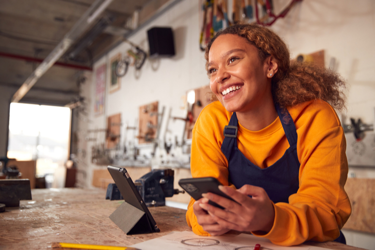Business Owner In Workshop Using Digital Tablet And Holding Mobile Phone
