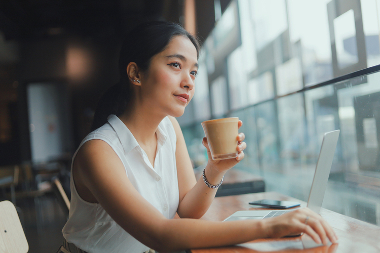 Asian woman using laptop while drinking coffee in cafe.