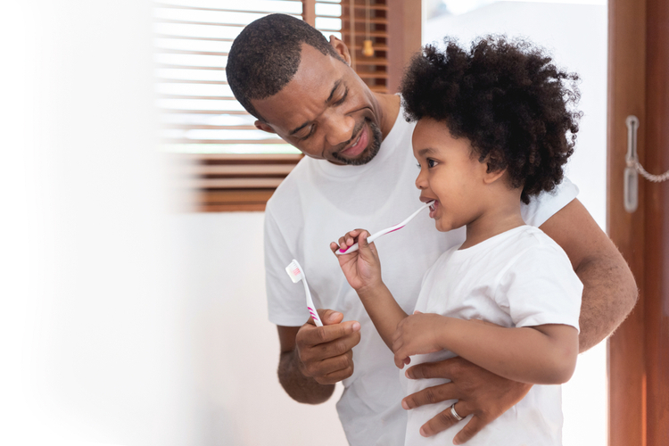 Dad teaching his son in white shirts brushing teeth in bathroom together.
