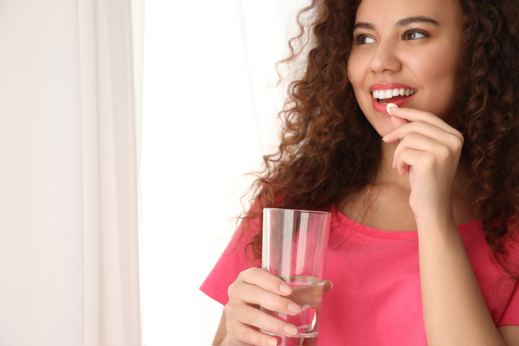 Woman with glass of water taking vitamin pill on light background.