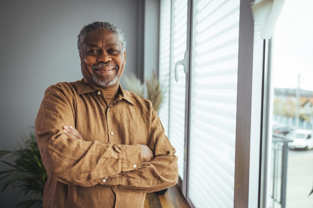 Black man in a button up shirt smiling by his window 