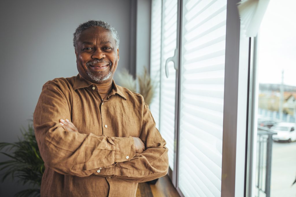 Black man in a button up shirt smiling by his window