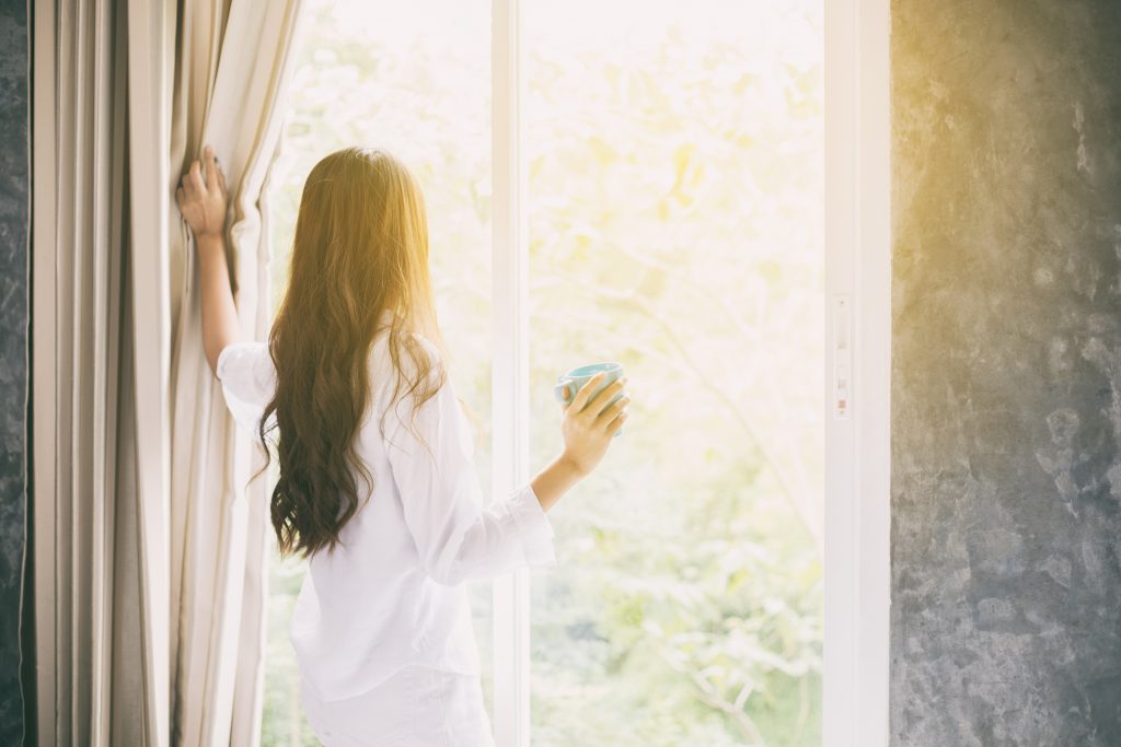 Woman holding a cup of coffee in her hand and opening up curtains to get sunlight