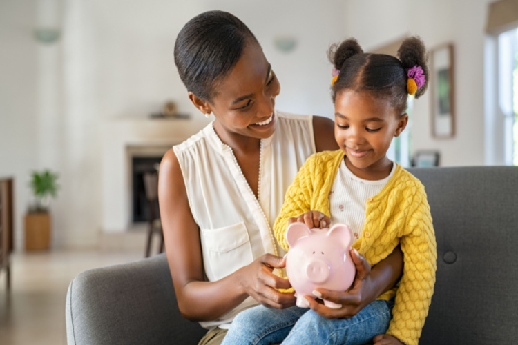 Mother helping daughter sitting on lap putting money in piggy bank.