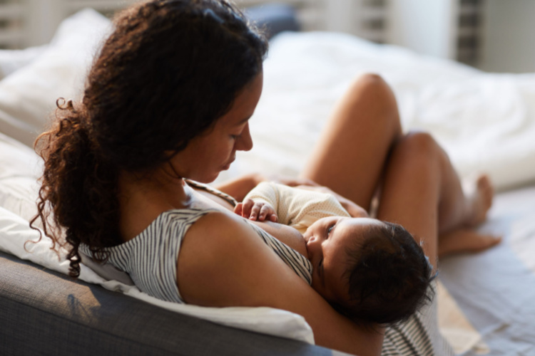 mother sitting on bed and leaning on headboard while feeding baby with breast