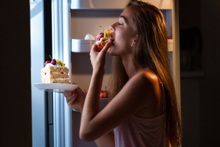 woman in pajamas eating sweet cakes at night near refrigerator.