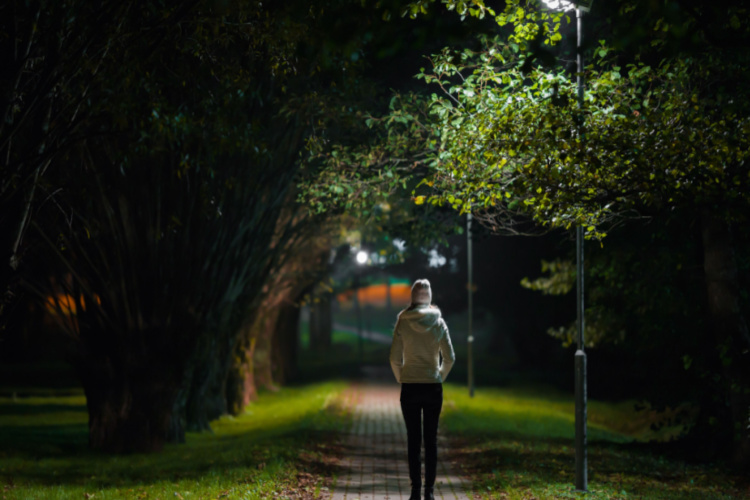 woman in white jacket walking on sidewalk through alley of trees under lamp light in autumn night.