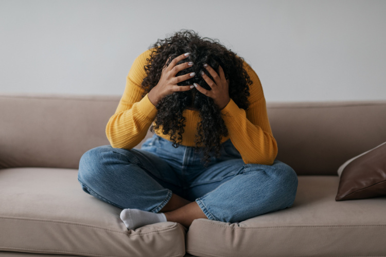 Black woman sitting cross legged on sofa with head in her hands at home.