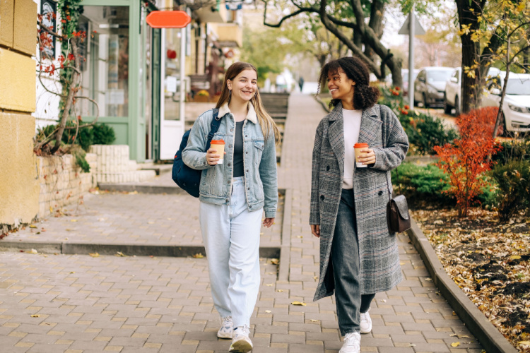 Two happy carefree multiracial girlfriends walking city street with take away coffee