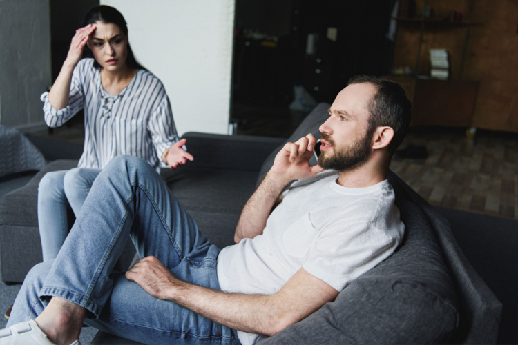 upset woman talking to her husband while he talking by phone on couch
